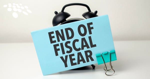 A black alarm clock holds a light blue note that reads "END OF FISCAL YEAR" in black text. The note is attached with a silver binder clip. The background is white.